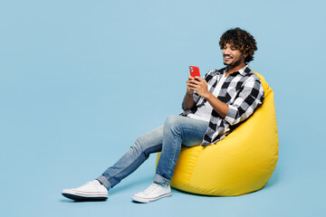 Full body young Indian man he wear shirt white t-shirt casual clothes sit in bag chair hold in hand use mobile cell phone isolated on plain pastel light blue cyan background studio. Lifestyle concept