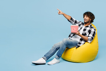 Full body young Indian man he wear shirt white t-shirt casual clothes sit in bag chair eat popcorn...