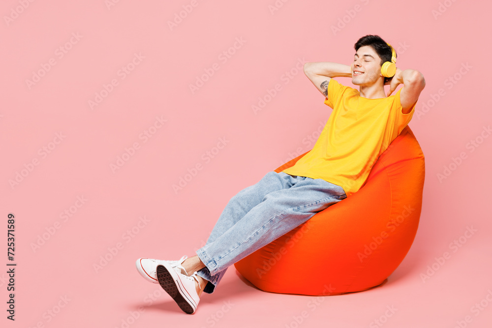 Wall mural Full body relaxed young man he wearing yellow t-shirt casual clothes sit in bag chair listen to music in headphones isolated on plain pastel light pink background studio portrait. Lifestyle concept.