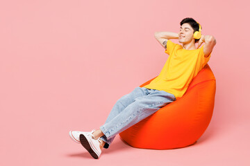 Full body relaxed young man he wearing yellow t-shirt casual clothes sit in bag chair listen to music in headphones isolated on plain pastel light pink background studio portrait. Lifestyle concept.