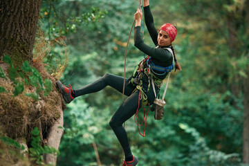 Looking to the side, hanging on the rope. Woman is doing climbing in the forest by the use of...