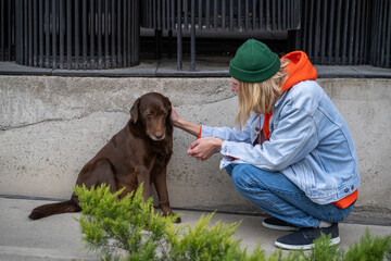 Long-haired stylish hipster guy sitting on haunches, stroking, caressing homeless stray dog in city...