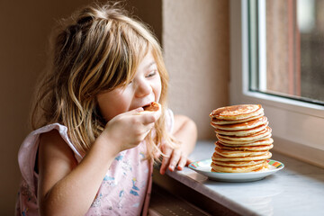Little happy preschool girl with a large stack of pancakes for breakfast. Positive child eating healthy homemade food in the morning. - obrazy, fototapety, plakaty