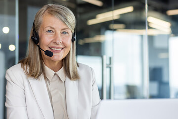 Portrait of mature lady in headset working by computer and smiling at camera on blurred background....