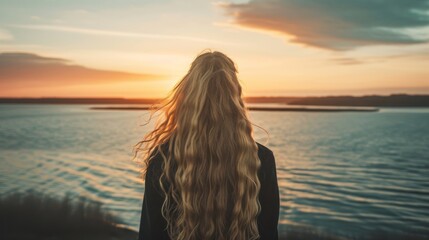 A woman with wavy, beautiful hair is standing in front of a body of water, watching the sunset and looking at the ocean waves.