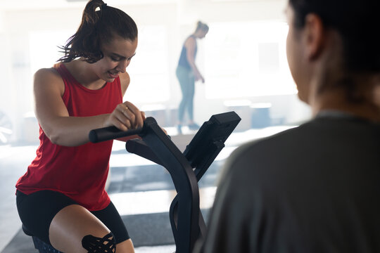 Caucasian woman motivating female friend exercising on elliptical trainer machine in gym