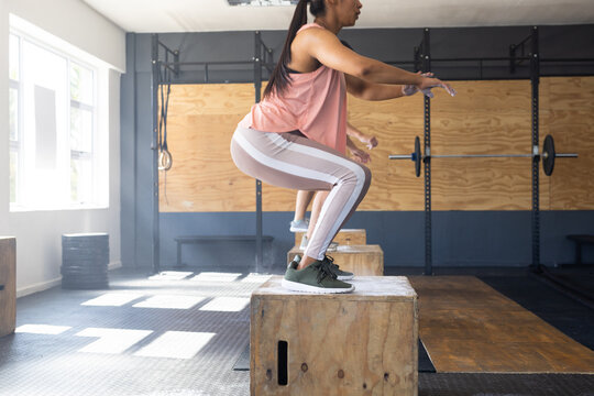 Side view of biracial young woman practicing squats on wooden plyo box in gym, copy space