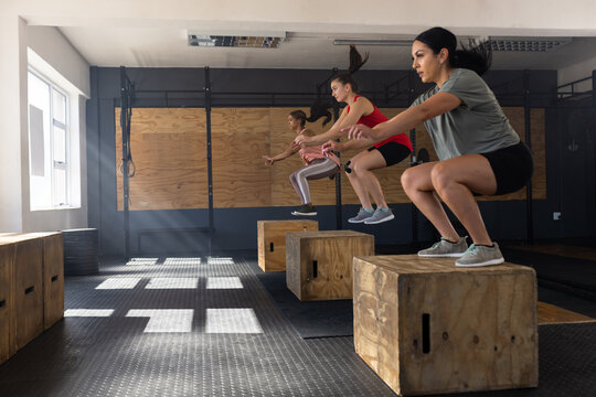 Side view of diverse women squatting and jumping on wooden plyo boxes in health club, copy space