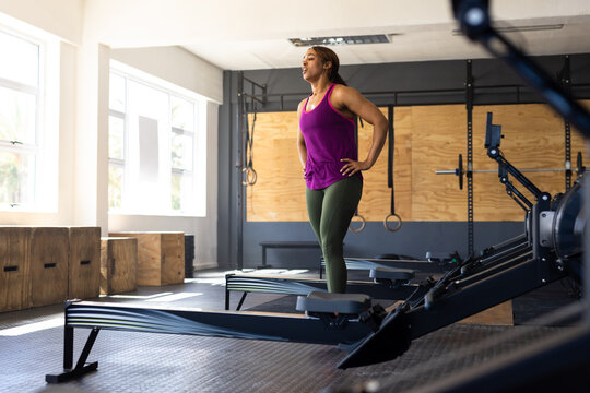 Exhausted biracial young woman with hands on hip standing by rowing machines in health club