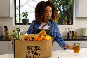 Happy biracial woman unpacking grocery shopping from recycled box in kitchen