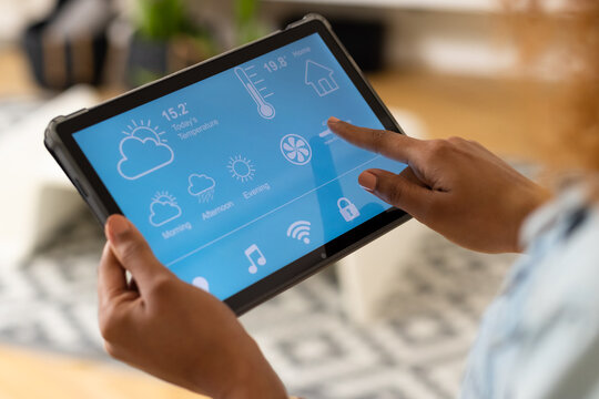 Hands of biracial woman in living room at home using tablet with smart home interface on screen