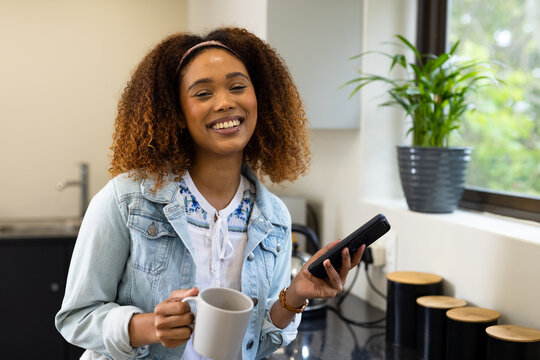 Portrait of happy biracial woman having coffee, smiling and using smartphone in kitchen at home