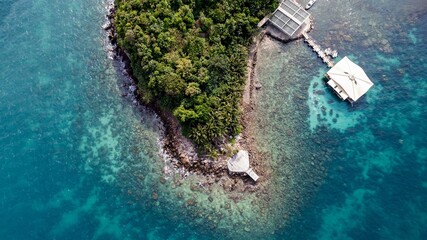 Aerial view of a tropical island with lush greenery surrounded by a crystal-clear coral sea, featuring a small pier and pavilion, ideal for travel or vacation concepts