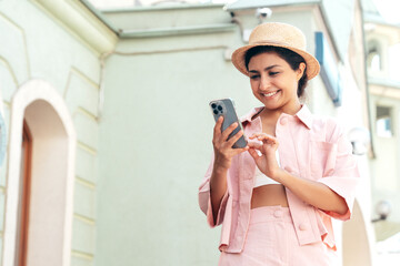 Young beautiful smiling hipster woman in trendy summer pink costume clothes. Carefree female posing in the street at sunny day. Positive model looks at cellphone screen, uses smartphone apps