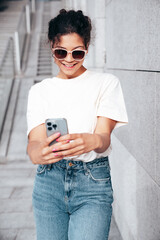 Young beautiful smiling hipster woman in trendy summer casual clothes. Carefree woman posing in the street. Positive model holds mobile phone, looks at cellphone screen, uses smartphone apps © halayalex