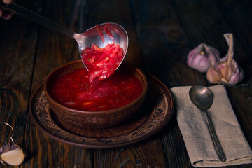 National Ukrainian borscht, earthenware, ladle pouring borscht into a plate, garlic, napkin and spoon on a wooden table