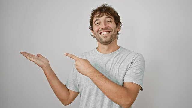 Amazed young man in t-shirt joyfully presents product, pointing with confident hand gesture on white isolated background. smiling heartily, hispanic guy creates a cheerful atmosphere.