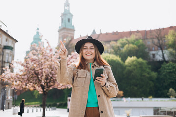 Attractive young female tourist in hat is exploring new city. Redhead 30s girl with smartphone pointing finger near Wawel castle, Cracow, Poland. Traveling Europe in spring