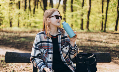 Happy blonde Woman in sport clothes having a halt after hiking. 30s Hiker drinking water from water bottle or hot drink from blue thermos
