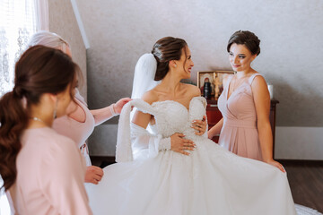 Wedding morning. Bridesmaids help put on the white wedding dress. A young woman is preparing to meet her groom and having fun with her friends