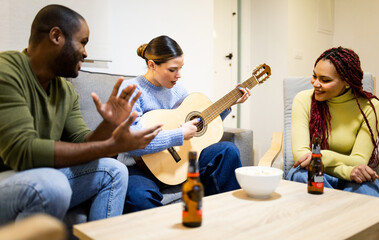 A group of 3 multiethnic young people are inside the living room of a house having fun with a...