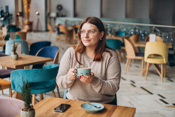 Attractive female in glasses with cute smile having a coffee while relax in in a break. Positive woman in casual wear smiling and resting with cup of hot drink in cozy cafe. Concept of easy breakfast