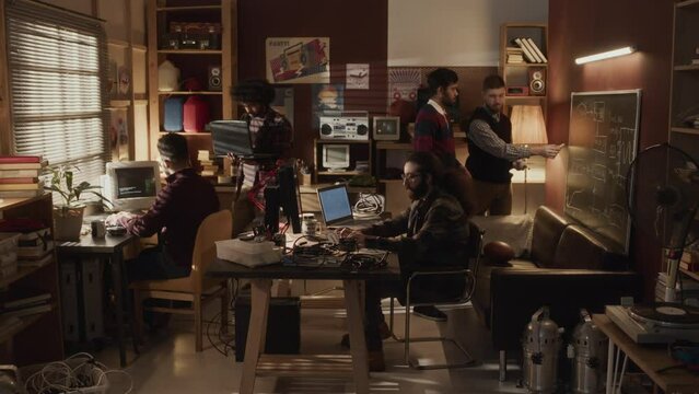 Full shot of group of multiethnic geeks interacting with each other in small room with warm dimmed light, working on software development and program coding