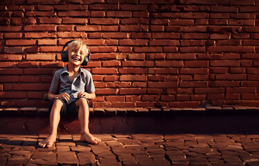 ein fröhlicher blonder Junge sitzt barfuß mit Kopfhörern lachend im sommer vor einer Mauer und...