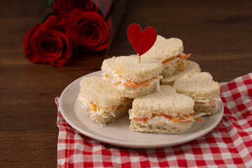 Heart shaped sandwiches made of salad sitting on a white dish on a red and white checkered cloth. There is a roses bouquet on the background. saint Valentines' food. Horizontal image