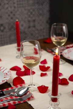 Dinner table set for two, with white wine and red roses' petals on the table. There is a lighted red candle. Vertical image