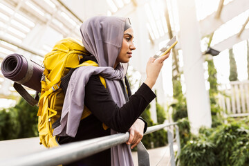 young veiled woman listening to an audio message on her cell phone while carrying her backpack and mat - obrazy, fototapety, plakaty