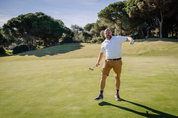 Sotogrante, Spain - January 25, 2024 - A golfer in a white shirt and tan shorts gestures with a putter on the green with trees in the background.