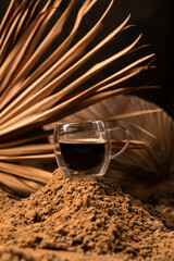 Transparent cup with coffee on the sand among palm leaves