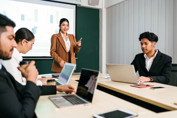 A cheerful and confident Asian businesswoman stands, present bar charts data from projector screen to her office colleagues. Asian business women leader role at the meeting.