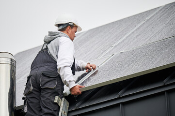 Man engineer mounting photovoltaic solar panels on roof of house. Technician in helmet installing...