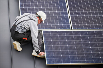 Man worker mounting photovoltaic solar panels on roof of house. Engineer in helmet installing solar module system with help of hex key. Concept of alternative, renewable energy.