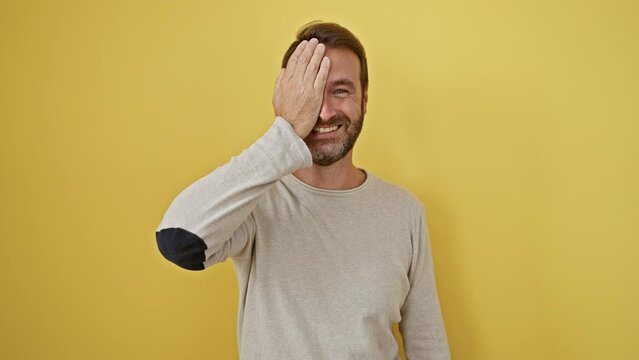 Handsome middle-aged hispanic man playfully covering one eye with hand, confident smile on his face against isolated yellow background. a picture of fun, cheeky portrait.