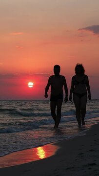 A man and a woman are walking along the seashore in front of a beautiful sunset.