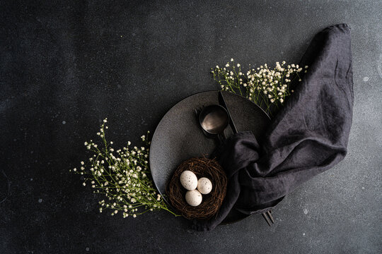 Overhead view of a black place setting with white gypsophila flowers and a bird's nest with Easter eggs on a black background