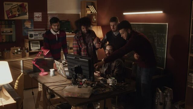 Lowkey shot of team of five multiracial male startupers having active discussions while standing around desk with computer, developing new software together in dark handmade old-fashioned office