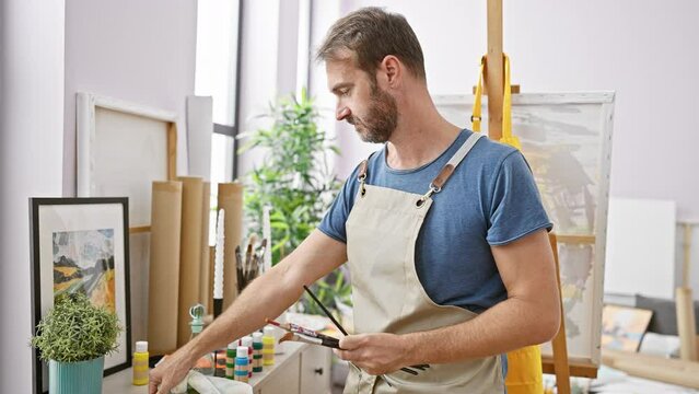 A focused bearded man in an apron paints in an artist's studio, surrounded by canvases, art supplies, and natural light.