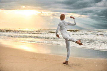 Young hairless ballerina with alopecia in white futuristic suit dancing on seashore at sunset sea, metaphoric surreal performance of bald pretty teenage girl exudes confidence, hope and unique beauty