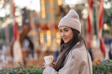 Smiling young woman holding a coffee cup in front of a carousel with bright lights. 