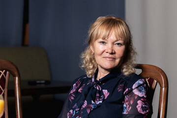 Portrait of happy 55 year old blonde woman sitting on chair indoors