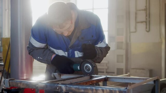 Slow motion video. Overall plan. Working man in overalls works with angle grinder in workshop. Mechanic removes old paint from metal surface. Cinematic light.