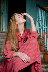 Tall slender caucasian young woman with blond hair in coral dress sitting on concrete stairs of old building with blue walls