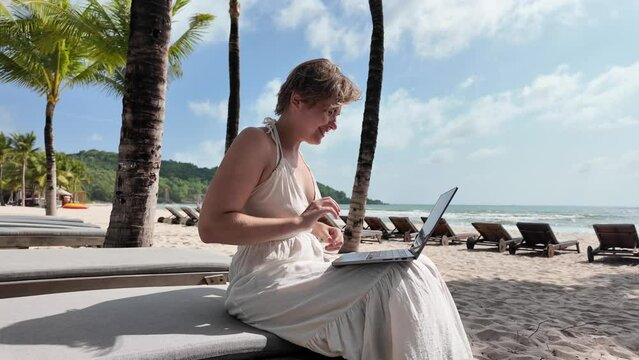 Adult woman working on a laptop at a tropical beach resort with palm trees and sun loungers in the background, concept of remote work or digital nomad lifestyle