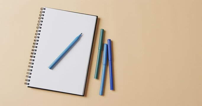 Overhead view of blue markers with notebook with copy space on beige background, in slow motion