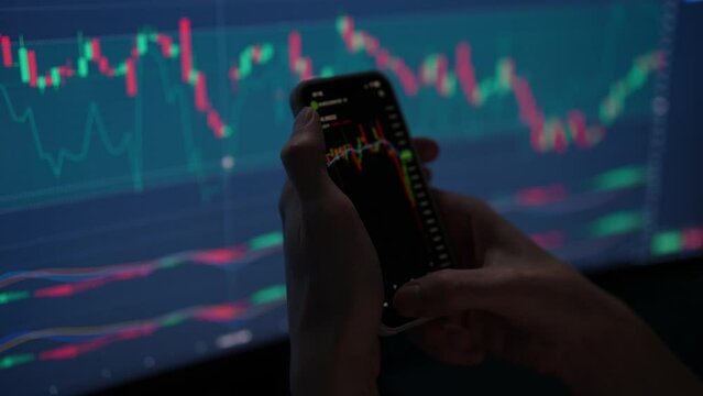 Closeup of trader analyzing bond market investments on smartphone with display chart of stocks or cryptocurrencies on background. Male investor holding using app cellphone for trading stock exchange