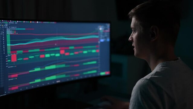 Focused financial male analyst using computer with display showing real-time stocks, exchange market charts. Remote working male stock trader making e-commerce investment sitting at desk in dark room.